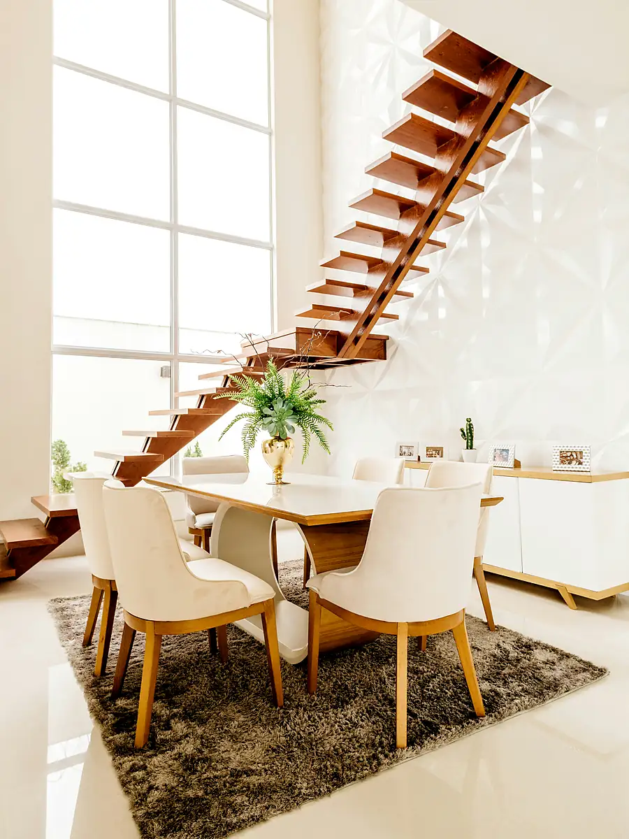 Escada de madeira e metal ao lado de uma mesa de jantar iluminada por janela alta